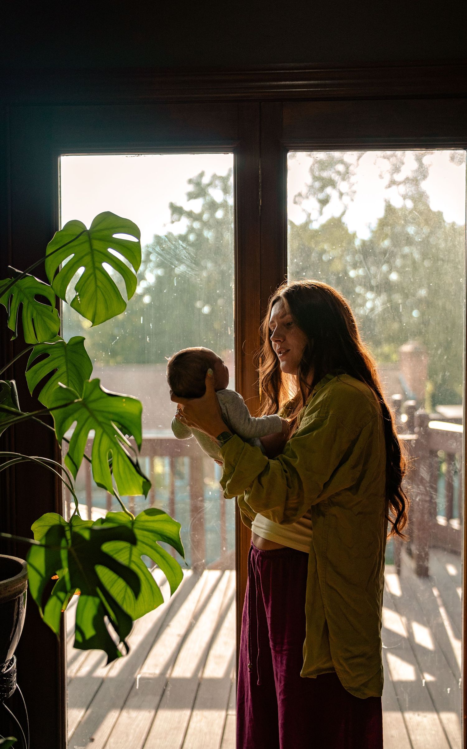A mother holds her newborn baby near a sunlit window with monstera plants, wearing a green top and red pants.