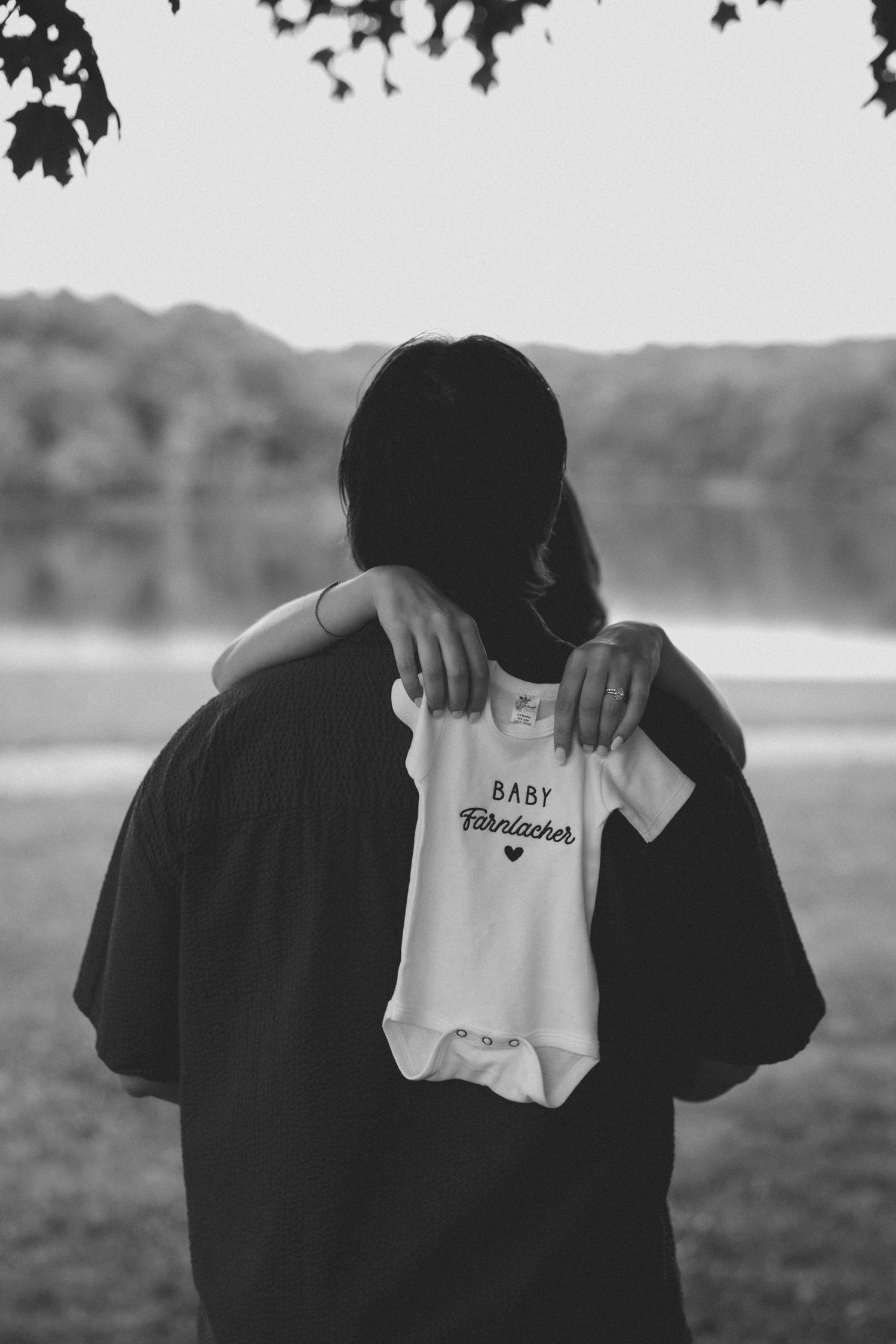 Black and white photo of a person holding a white baby onesie reading Baby Fairlacher outdoors near a lake.