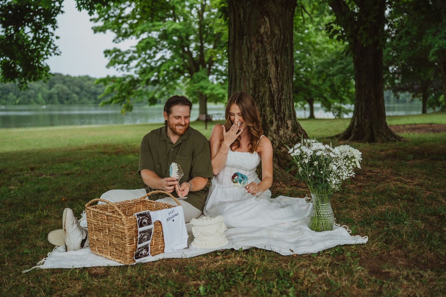 Couple sharing a romantic outdoor picnic under a tree by a lake with a wicker basket and white wildflowers.