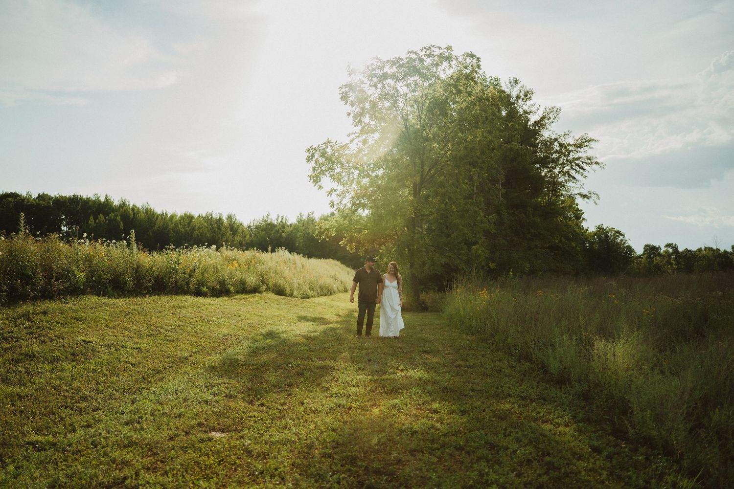 Couple walking hand in hand through a sunlit green meadow with tall trees glowing in golden backlight.