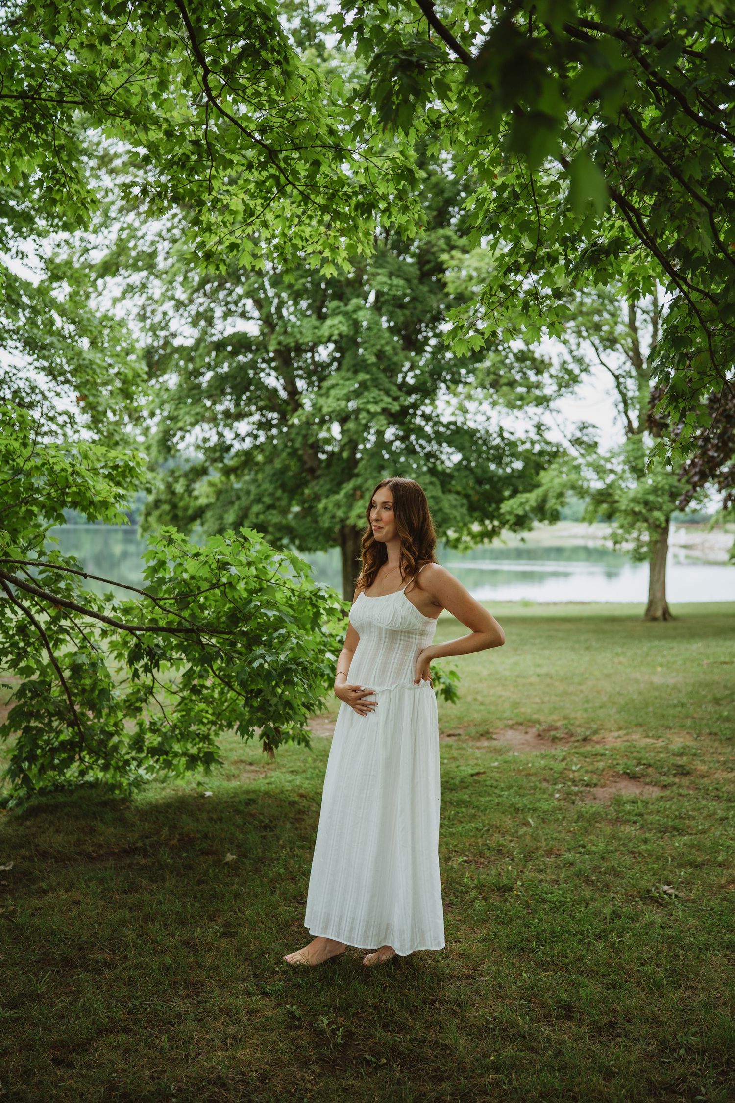 Pregnant woman in white maxi dress standing under lush green trees near a serene lake.