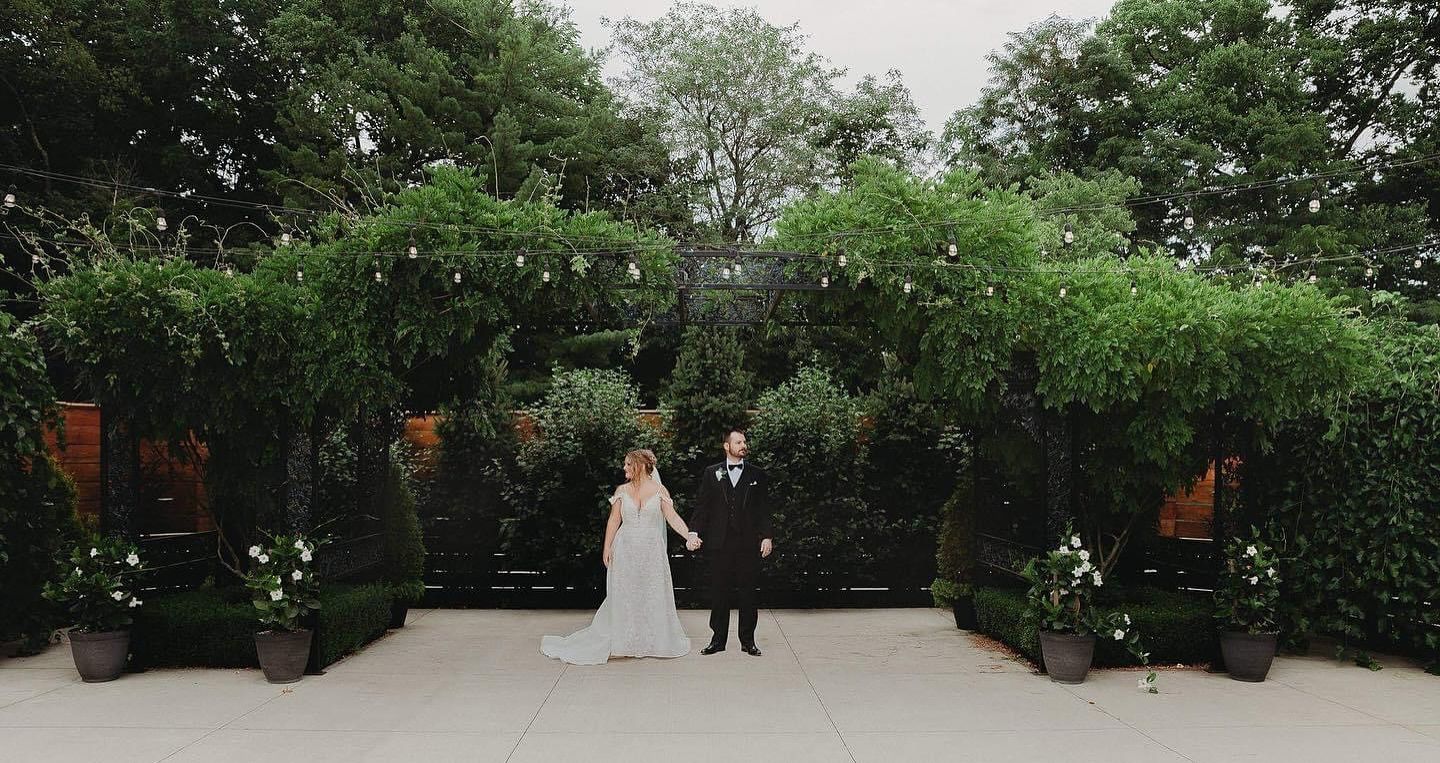 A couple stands under a romantic garden archway adorned with string lights and greenery at an outdoor wedding venue.