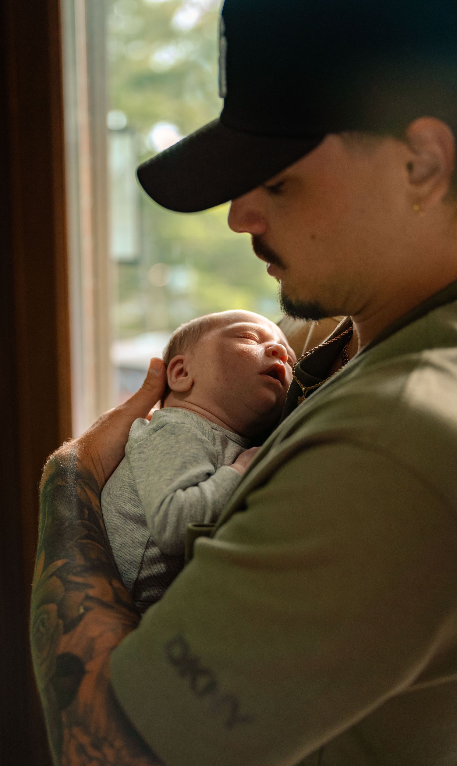 Tattooed father in black cap tenderly holds sleeping newborn baby near a sunlit window.