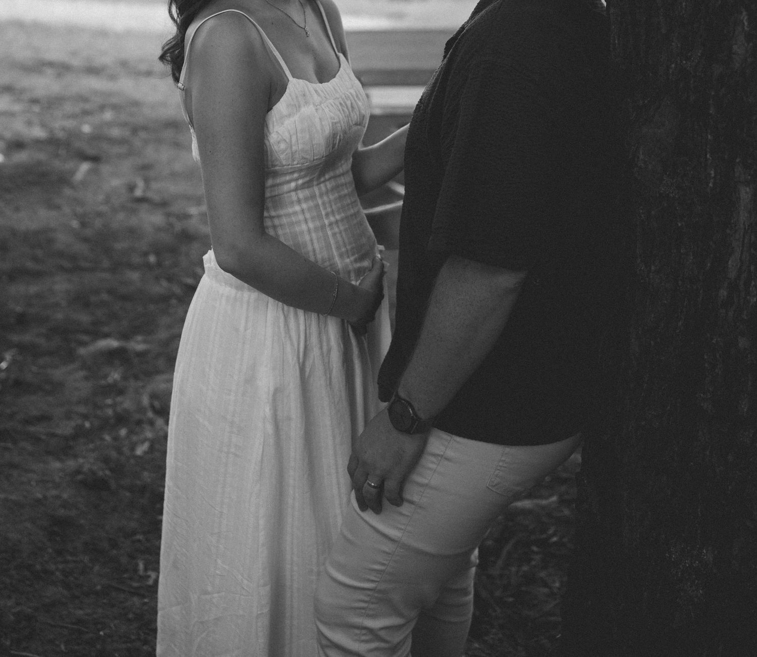Black and white photo of a couple holding hands near a tree, woman in white maxi dress.