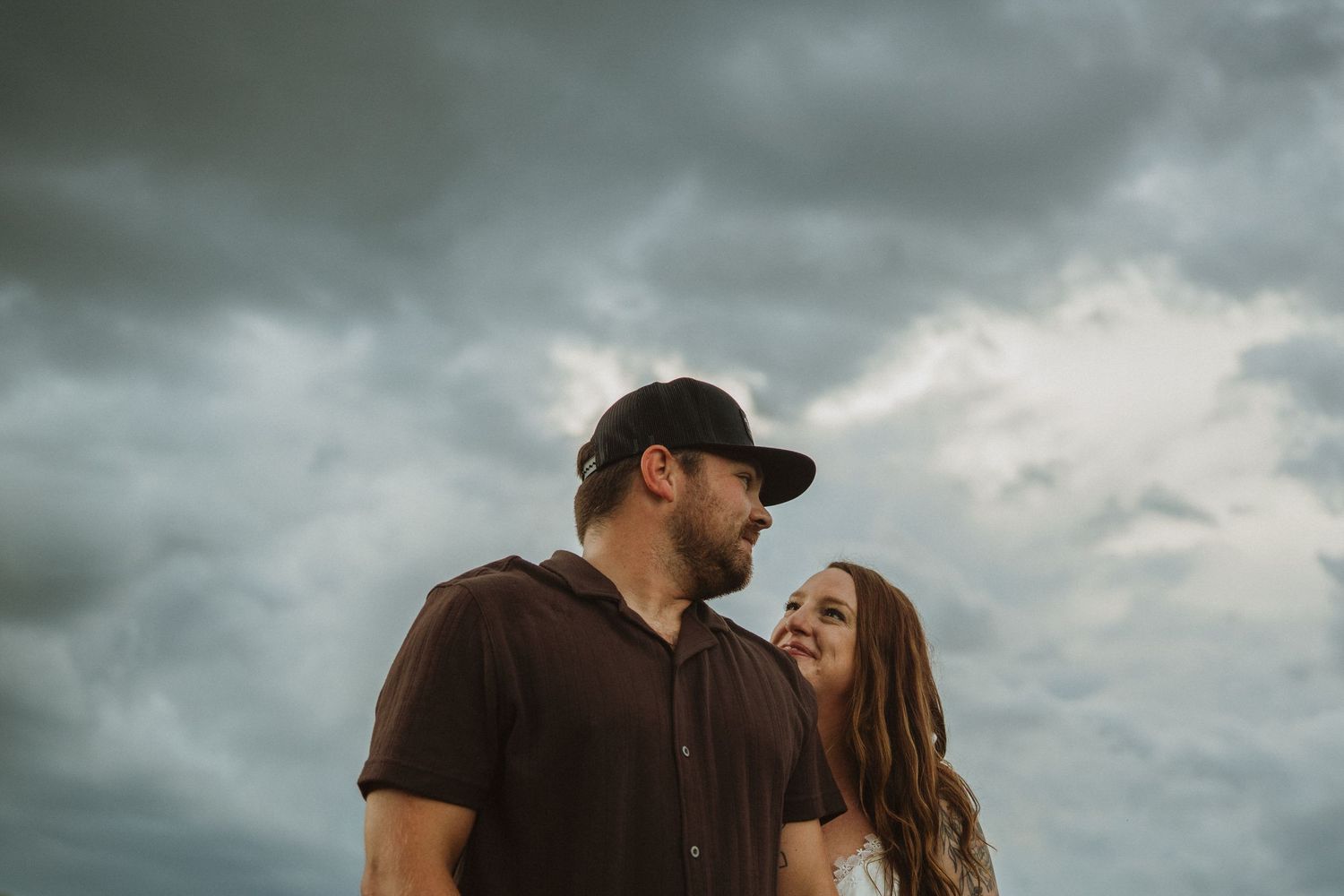 A couple stands close together under dramatic stormy clouds, the man in a cap and brown shirt, woman smiling softly beside him.