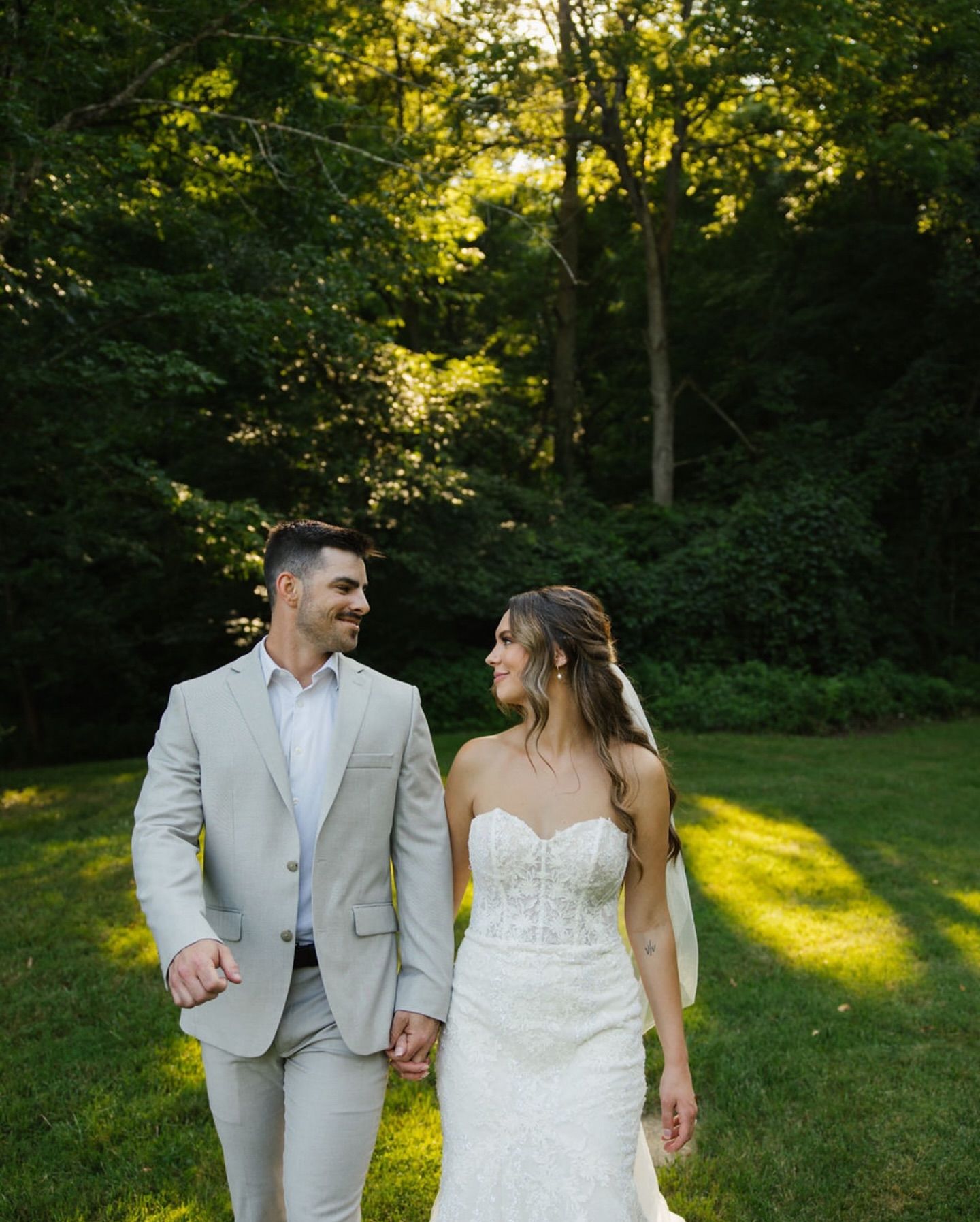 A couple walks hand in hand through a sunlit wooded area wearing wedding attire on their special day.