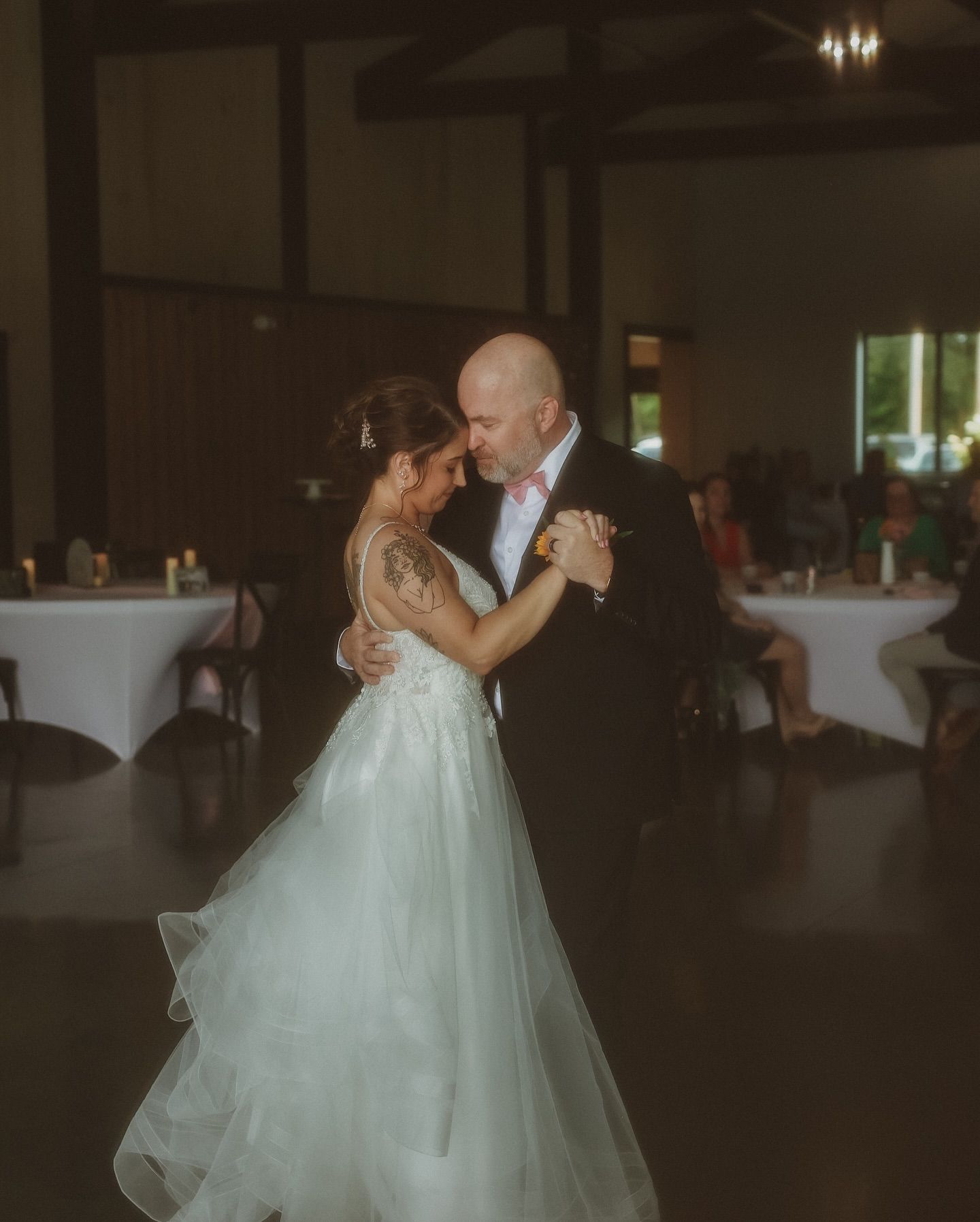 A couple shares an intimate dance moment in a dimly lit reception hall with white table settings in the background.