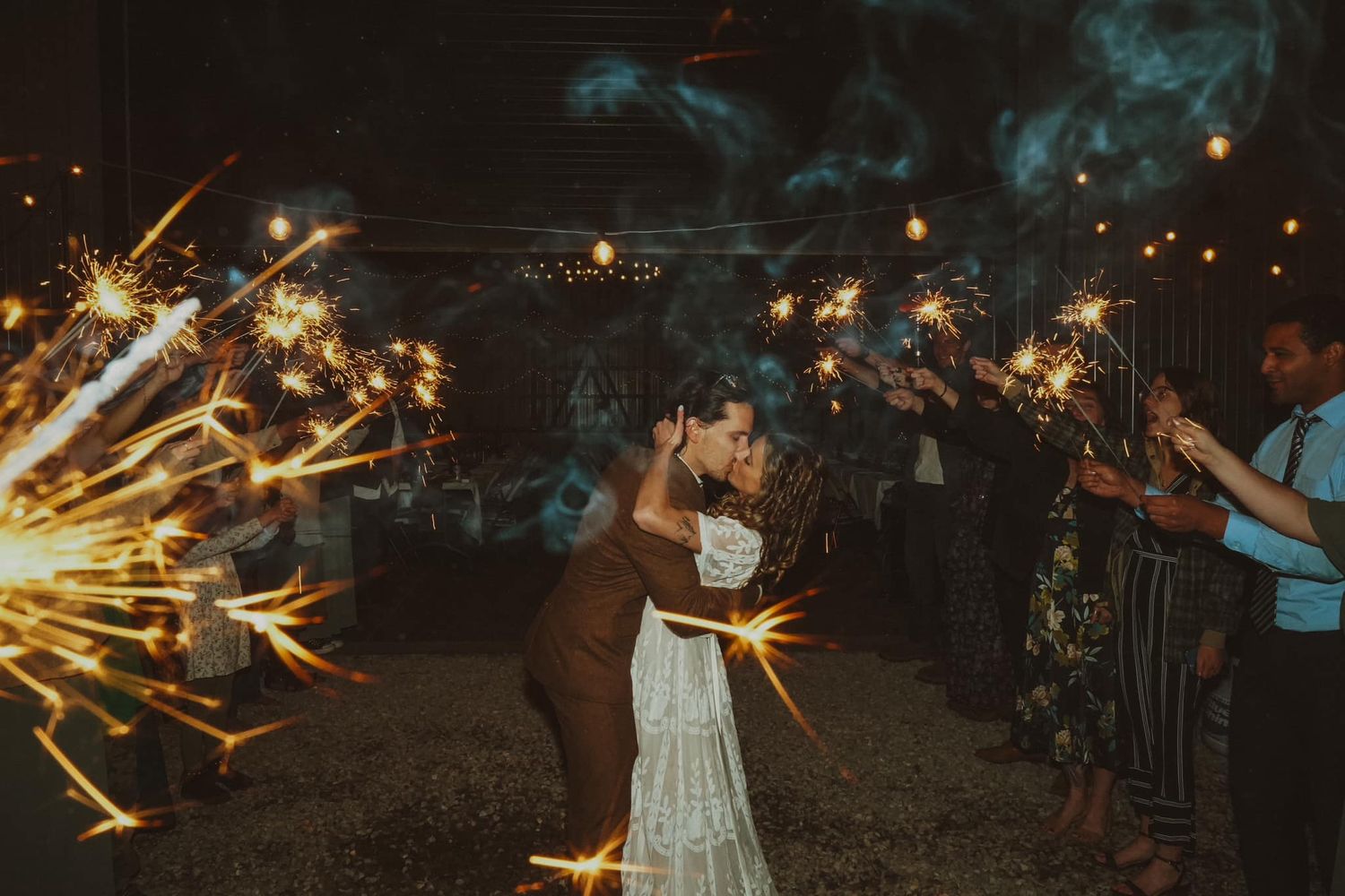 Wedding couple shares romantic kiss during sparkler exit with guests holding sparklers creating magical nighttime atmosphere.