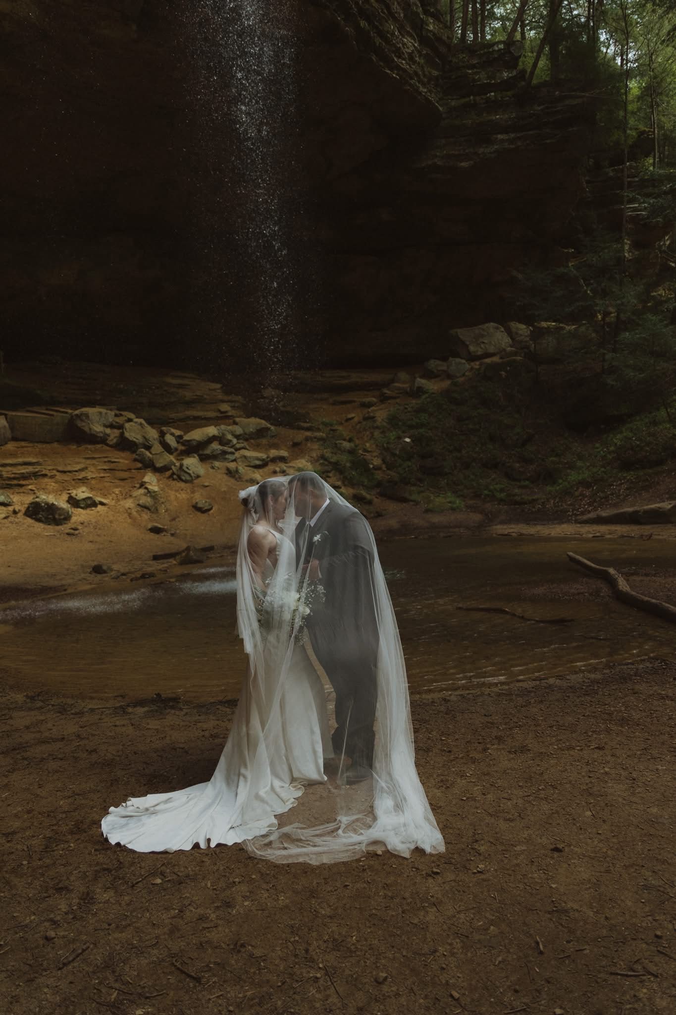 A romantic forest wedding photo shoot with a flowing white veil and formal attire against a dark wooded backdrop.
