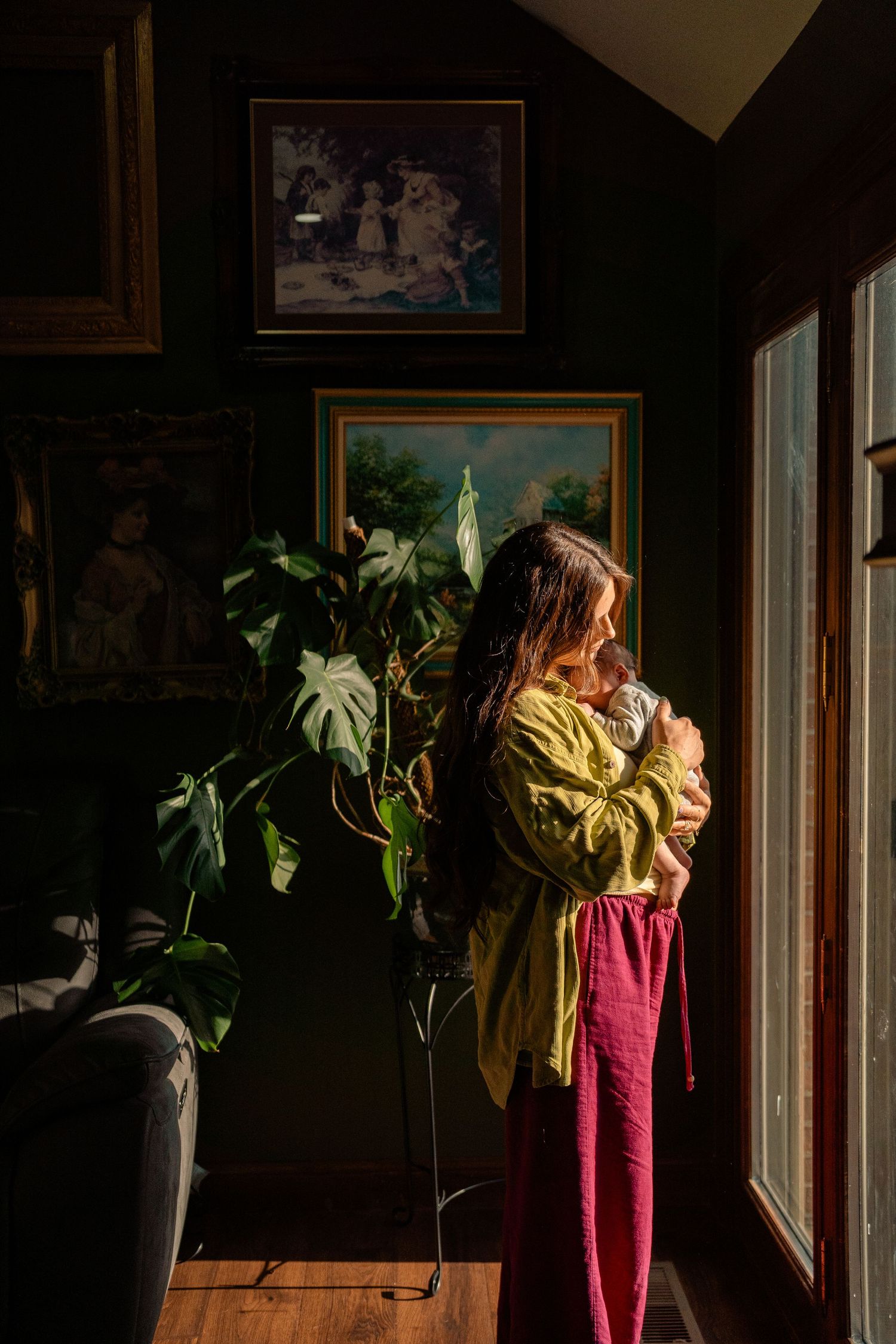 Mother in yellow top and burgundy pants holds newborn near sunlit window with dark walls and framed artwork.