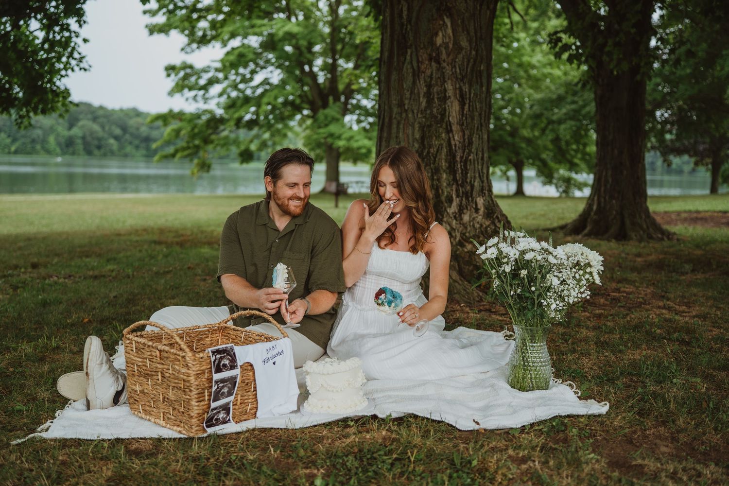 Couple enjoying a romantic outdoor picnic under large trees near a lake with a wicker basket and wildflowers.