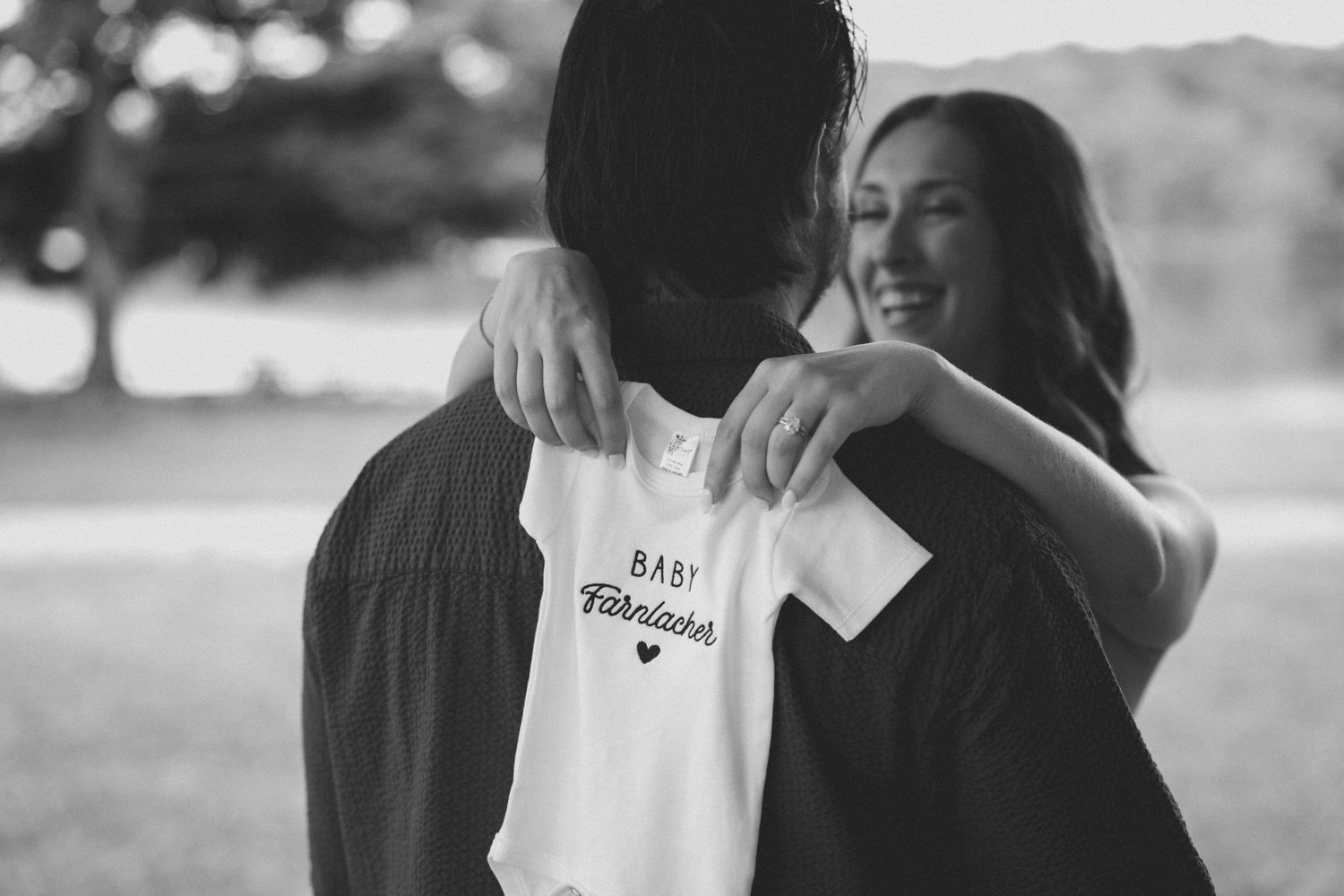 Black and white photo of couple embracing outdoors while woman holds a Baby Farbischer onesie.