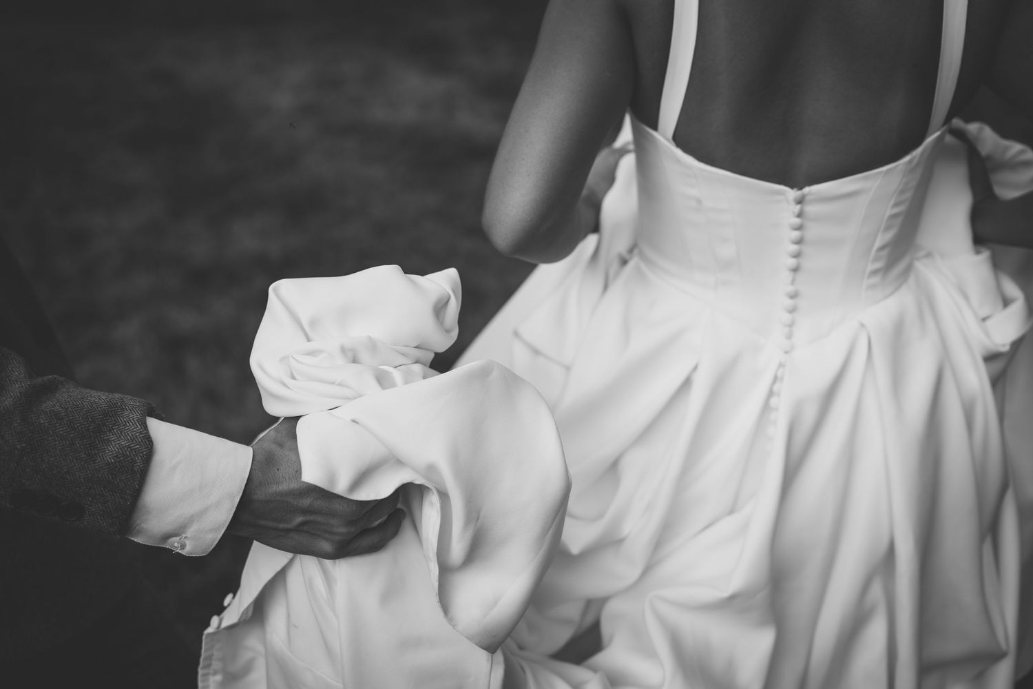 Black and white photo of a bride in a white button-back wedding gown being helped with her dress train.