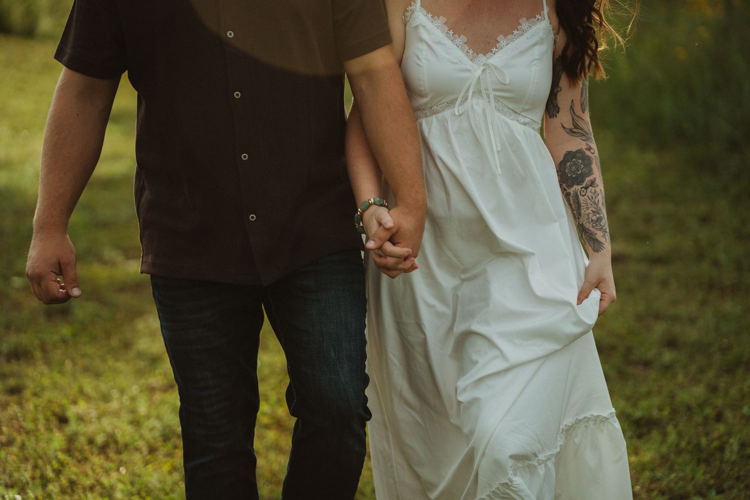 Couple holding hands walking outdoors, woman in white lace wedding dress with tattoos, man in dark shirt and jeans.