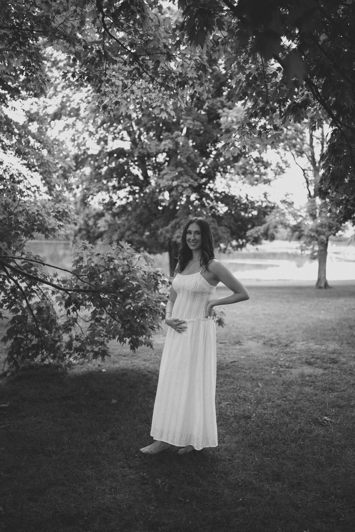 Pregnant woman in white maxi dress smiling outdoors in park under trees, black and white photo.