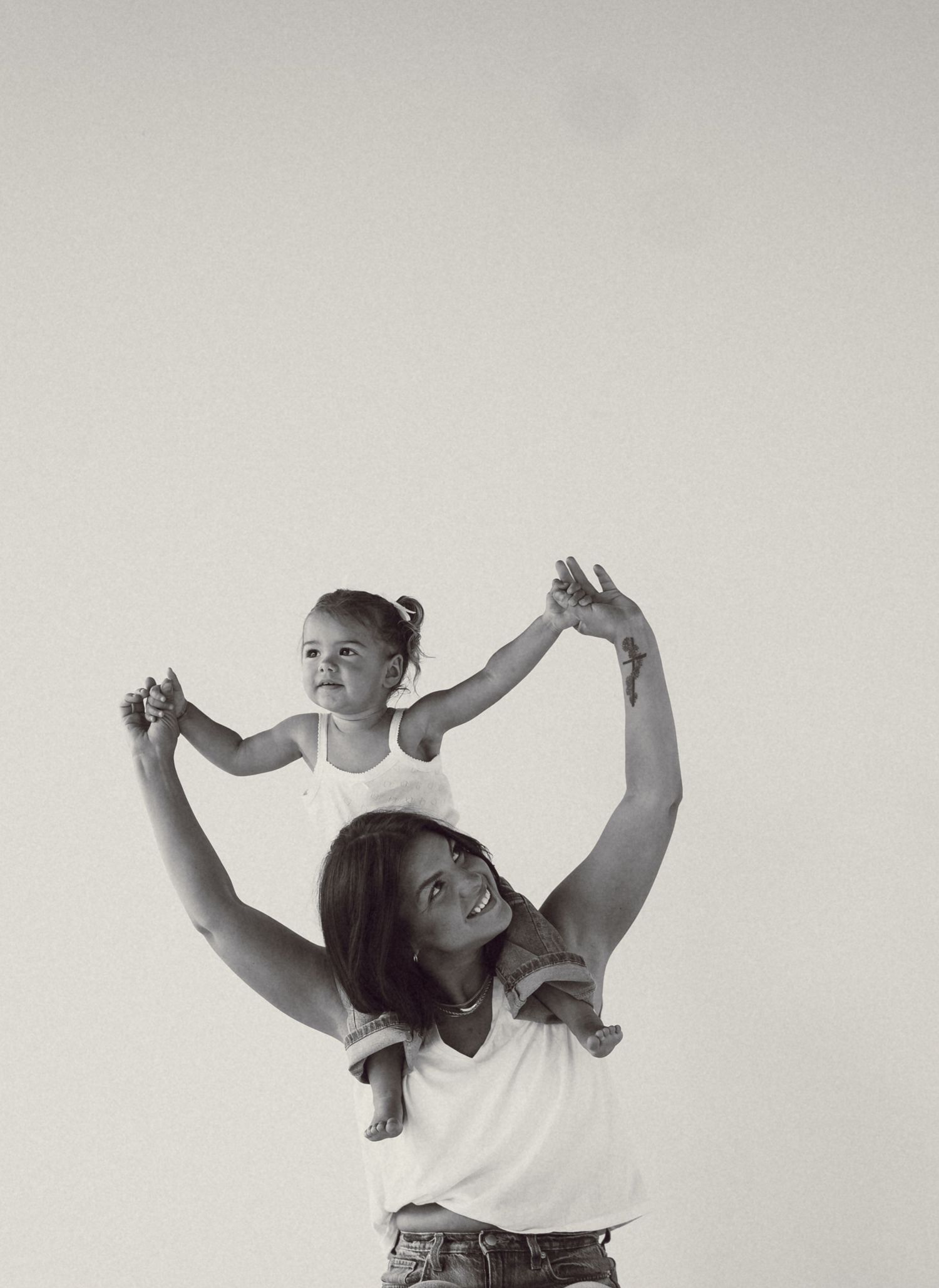 Black and white photo of a smiling woman holding a toddler on her shoulders, both with arms raised joyfully.