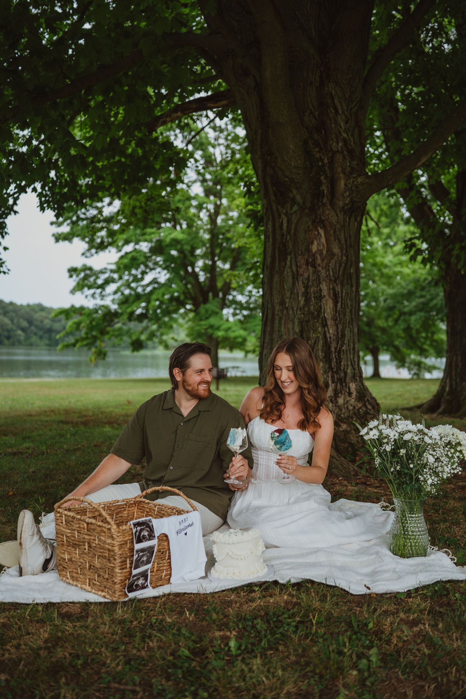 Couple sharing a romantic picnic under a large tree by a lake, with a wicker basket and wildflowers nearby.