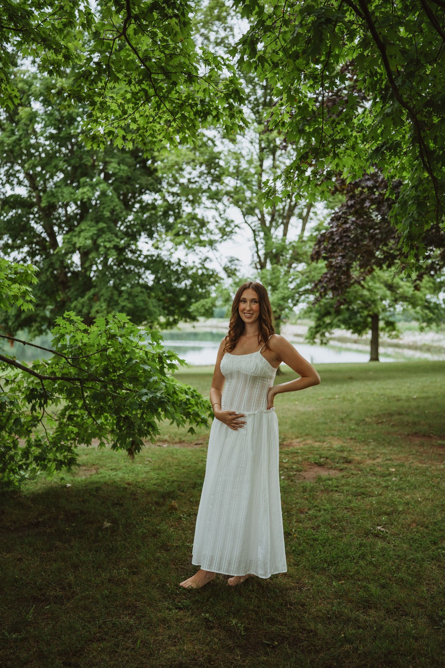 Smiling pregnant woman in white sundress standing in lush green park near a lake.