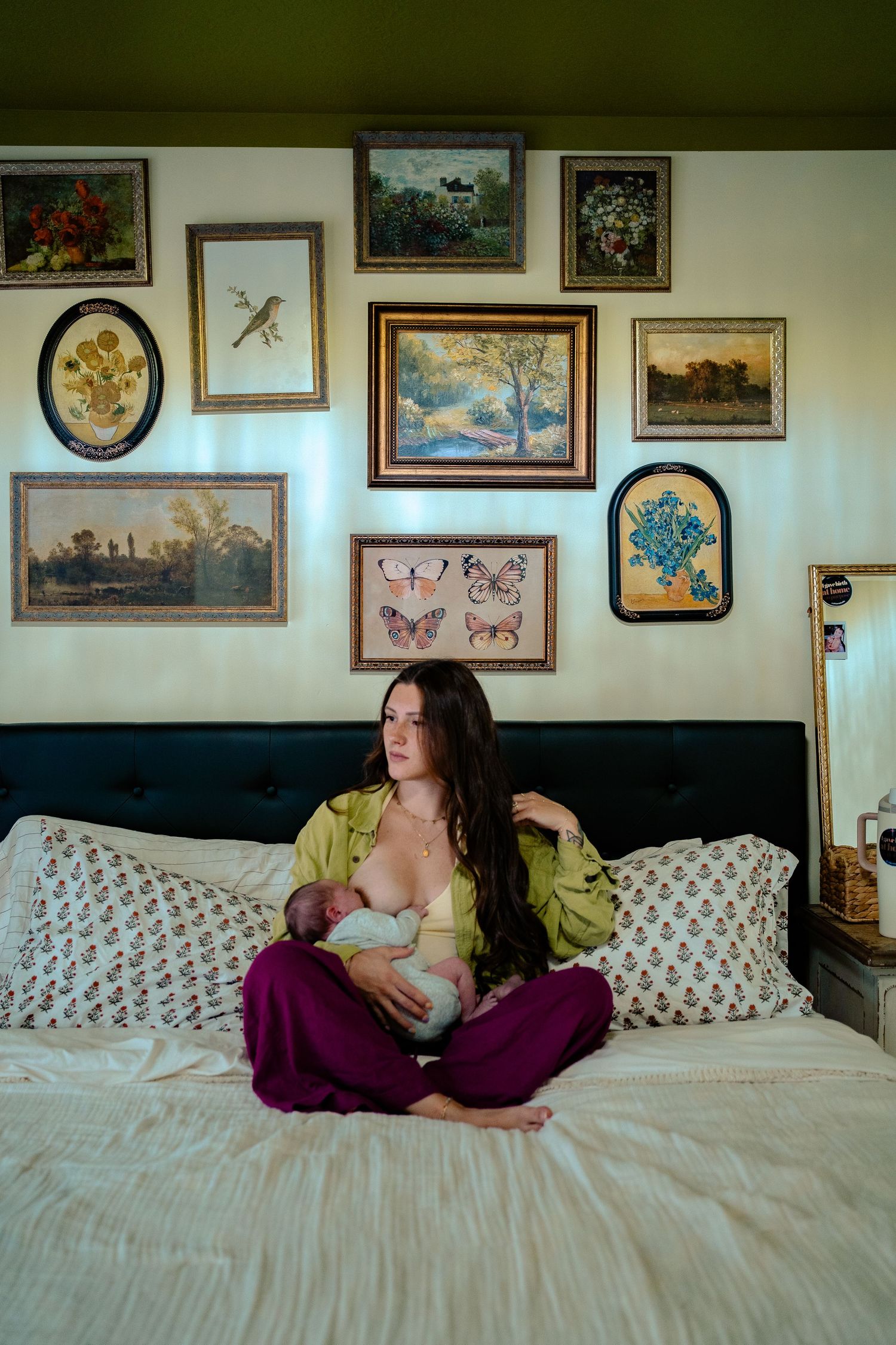 New mother in green silk top breastfeeds newborn on bed surrounded by eclectic gallery wall.