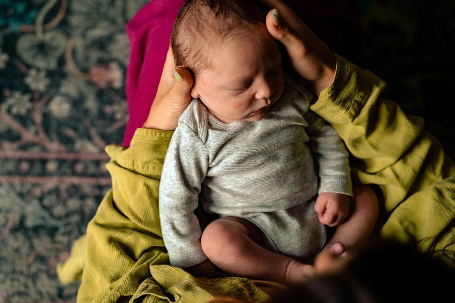 Newborn baby sleeping peacefully in parent's arms, dressed in gray onesie, with warm golden light.