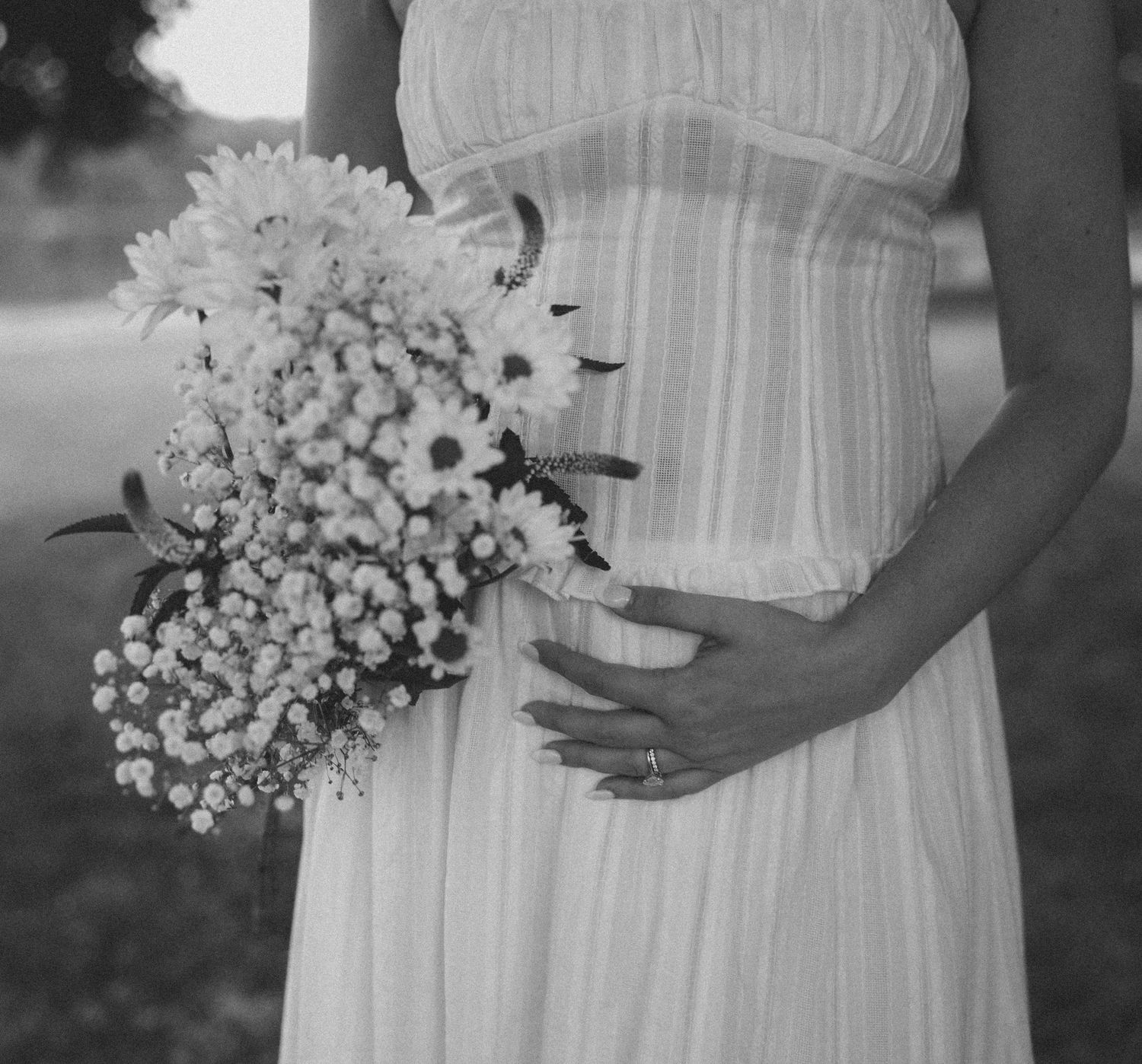 Black and white maternity photo of a pregnant woman in a white dress holding a wildflower bouquet.