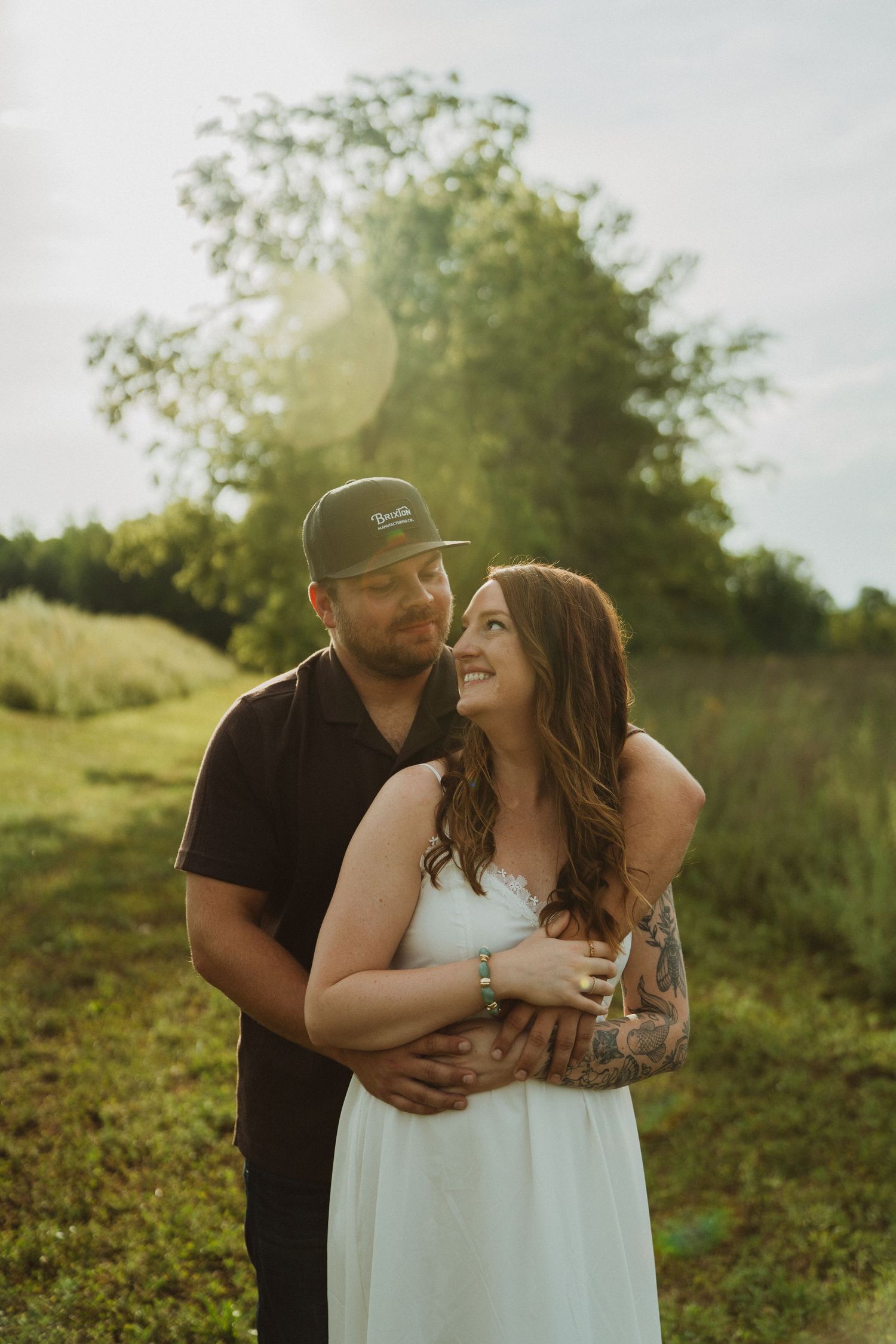Couple embracing in golden hour sunlight in a green field, woman in white dress smiling up at man in black polo and cap.