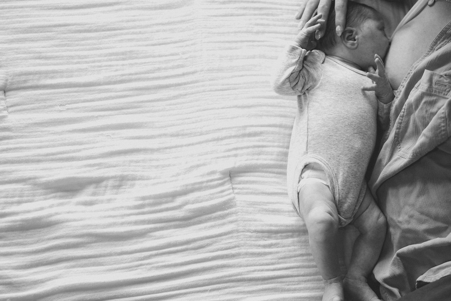 Black and white image of newborn baby in white onesie sleeping on rumpled bed.