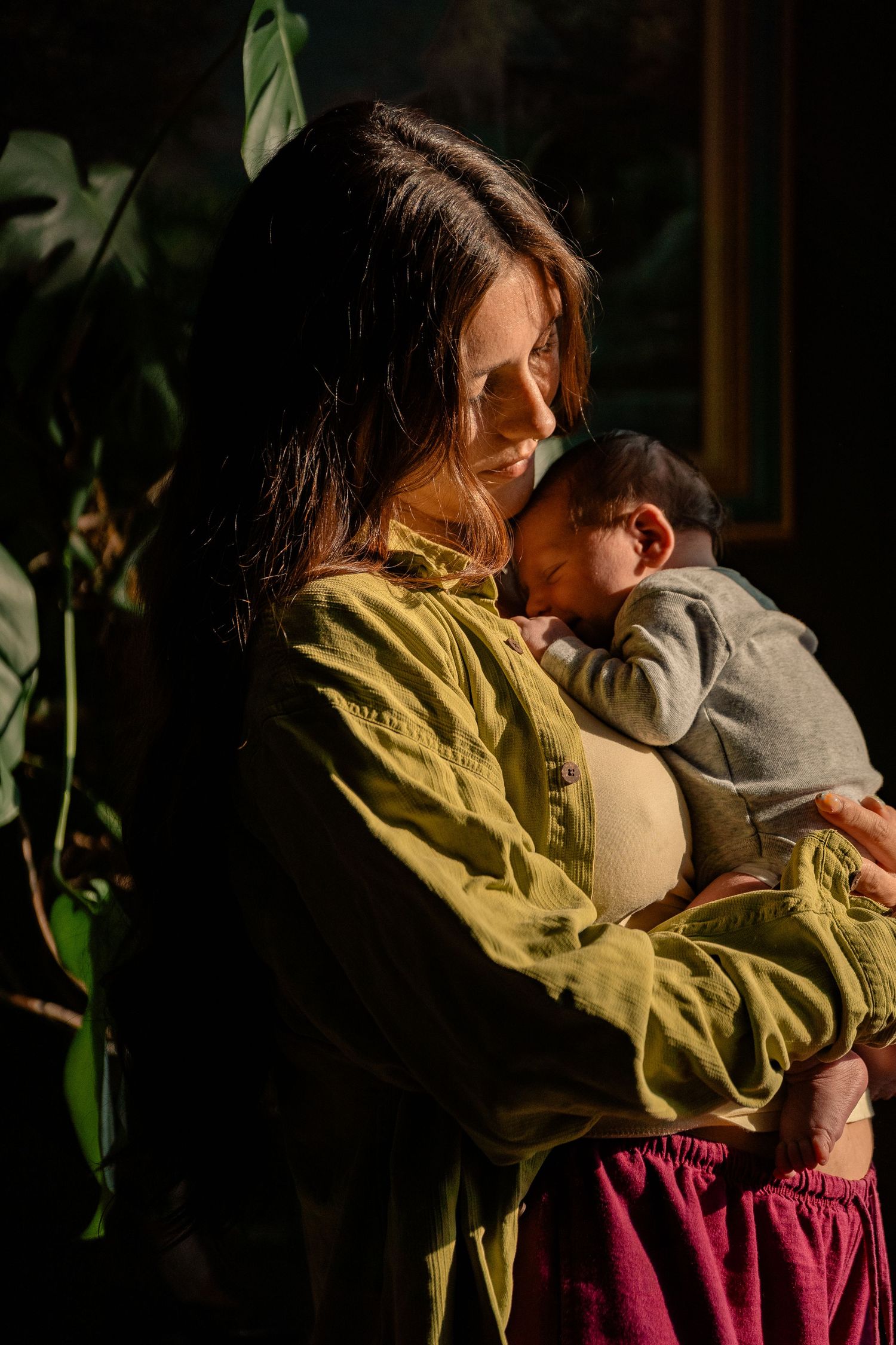 New mother in olive green shirt tenderly cradles sleeping newborn baby in golden natural light.