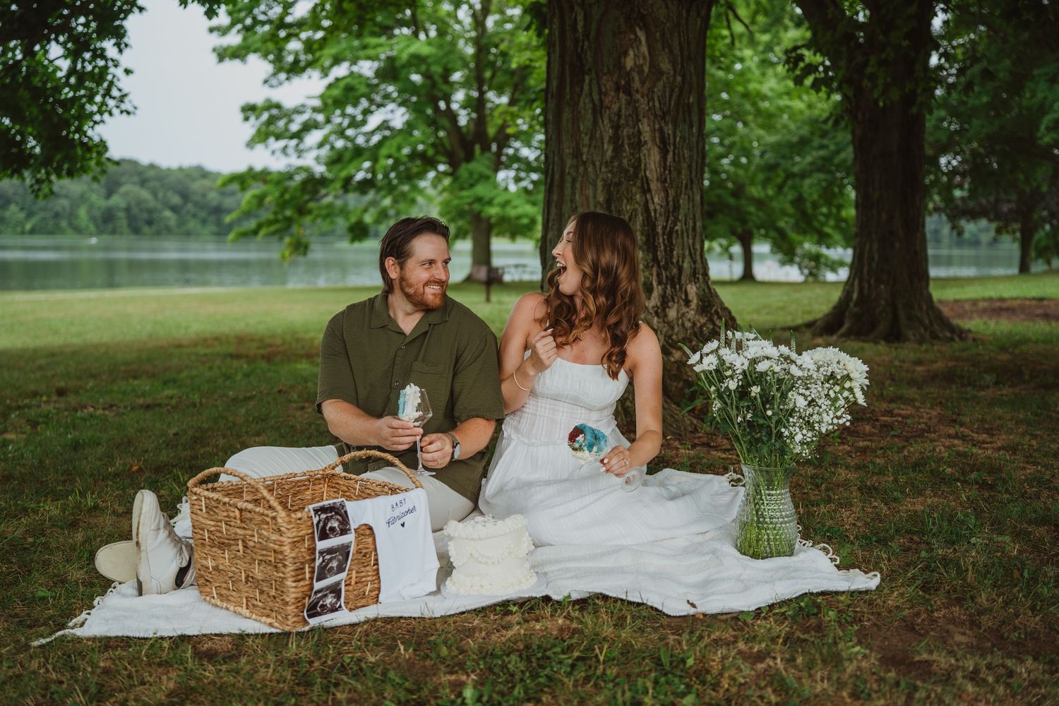Couple sharing a romantic picnic under trees by a lake, sitting on white blanket with wicker basket and wildflowers.