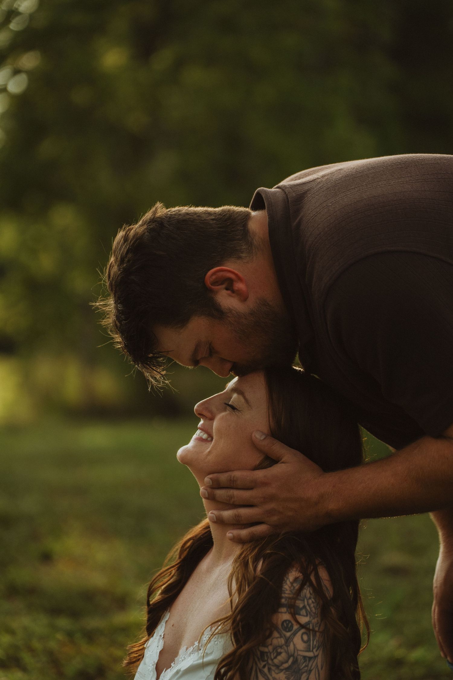 Man tenderly kissing tattooed woman's forehead in golden sunlight, romantic couple portrait in lush green outdoor setting.