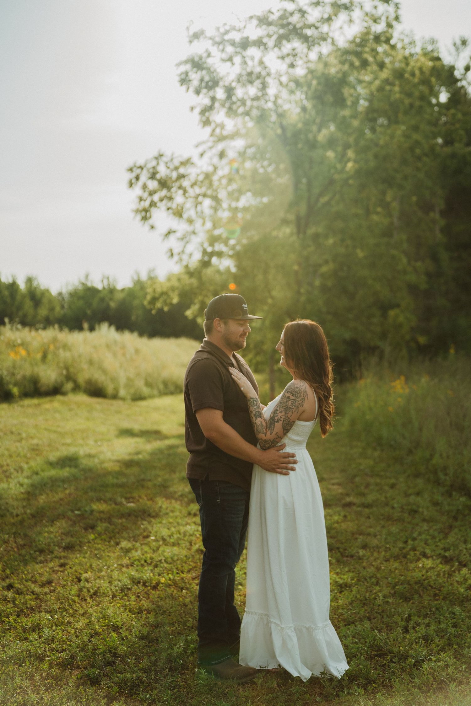 Couple embracing in a sunlit field, woman in white dress with tattoos, man in dark shirt and cap, lush green trees behind.