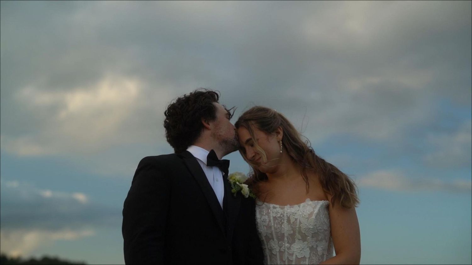 A groom in a black tuxedo kisses his bride in a white strapless gown outdoors under a cloudy blue sky.