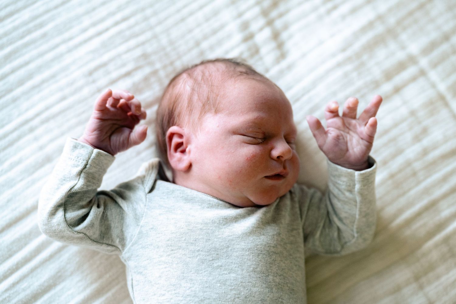 Peaceful newborn baby in gray onesie sleeping with arms raised on white bedding.