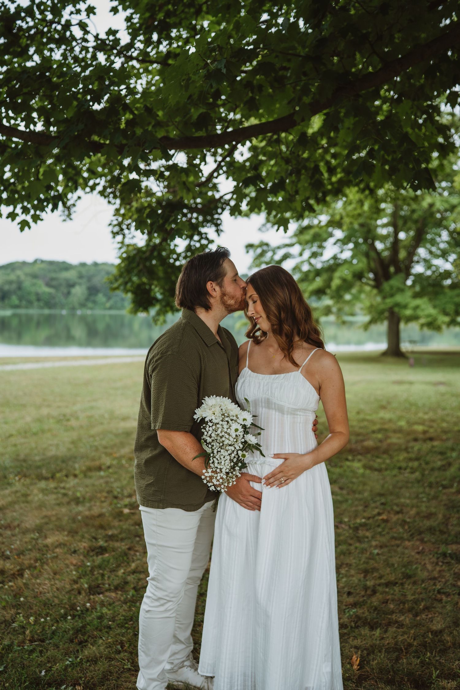 Couple kissing under a large tree by a lake during a maternity photoshoot, holding white flowers.