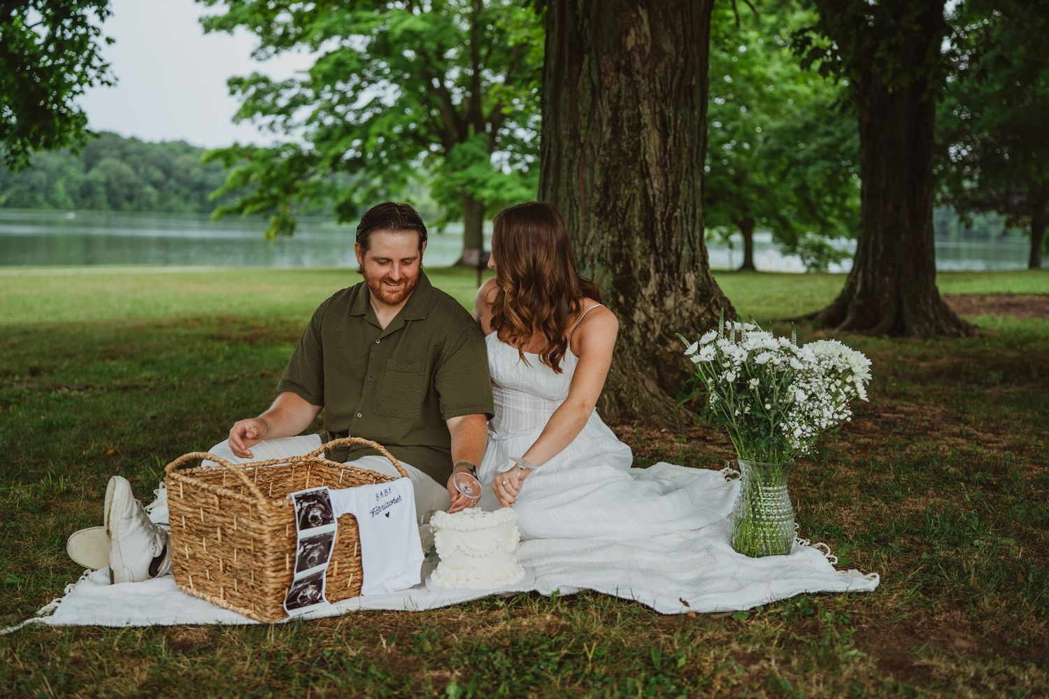 Couple sharing a romantic picnic under trees near a lake, looking at ultrasound photos in a wicker basket.