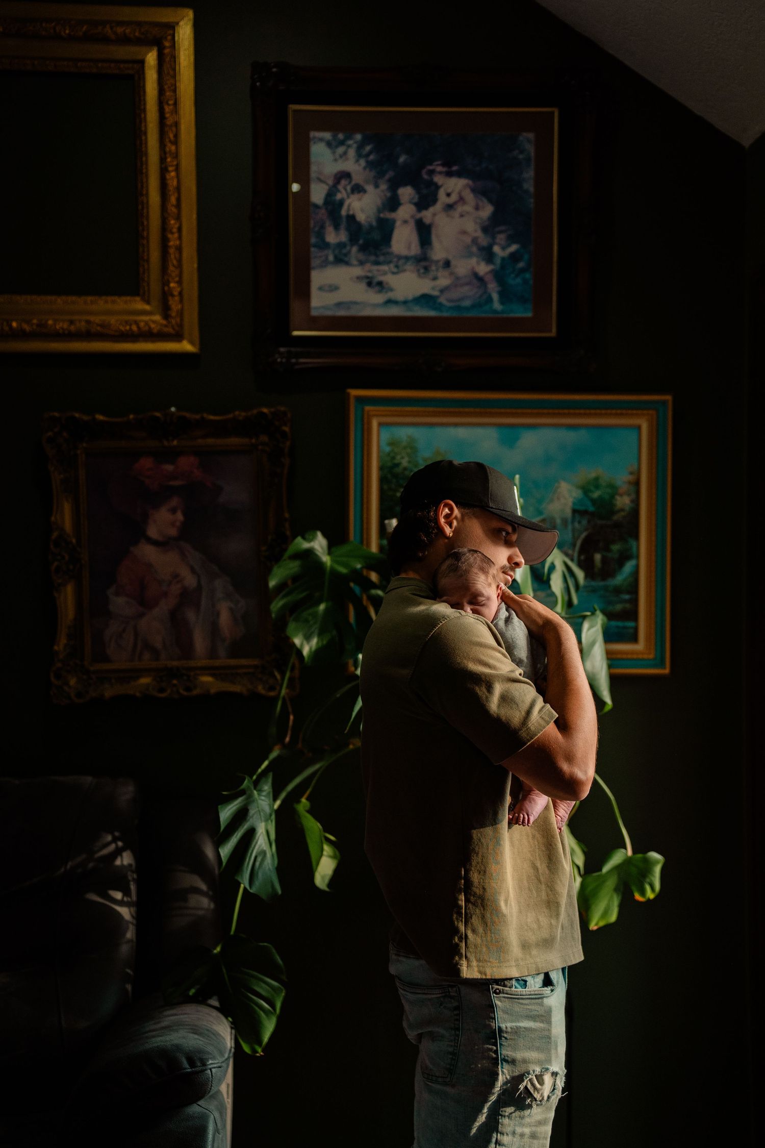 Father in cap gently holds newborn baby against his chest in a dark room with vintage artwork.
