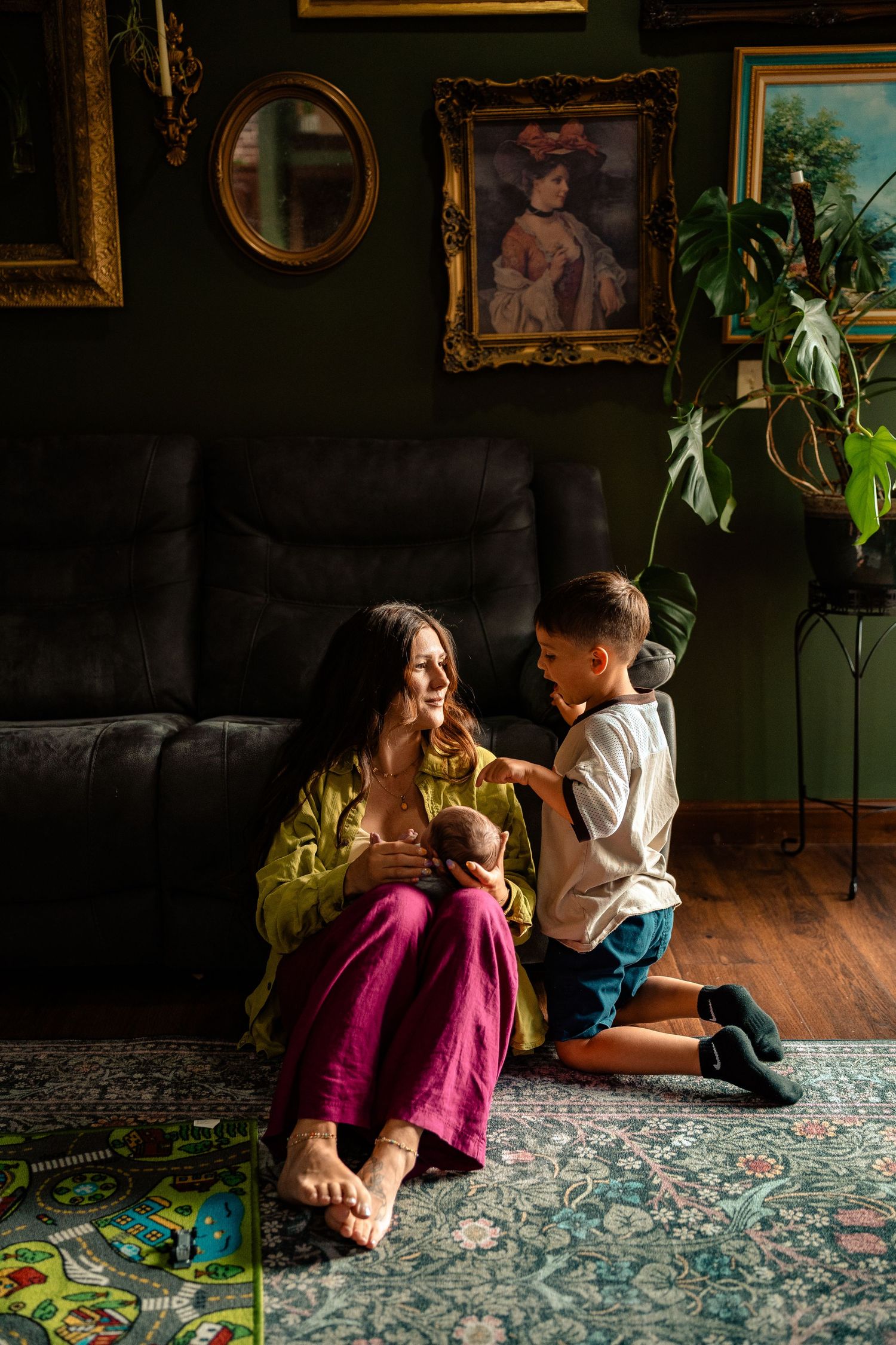 Mother sits on floor holding newborn while young son kneels beside her in a dark moody living room.