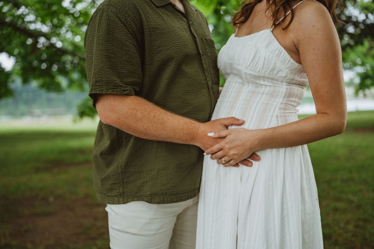 Close-up of couple holding pregnant belly together outdoors under green tree during maternity photoshoot.