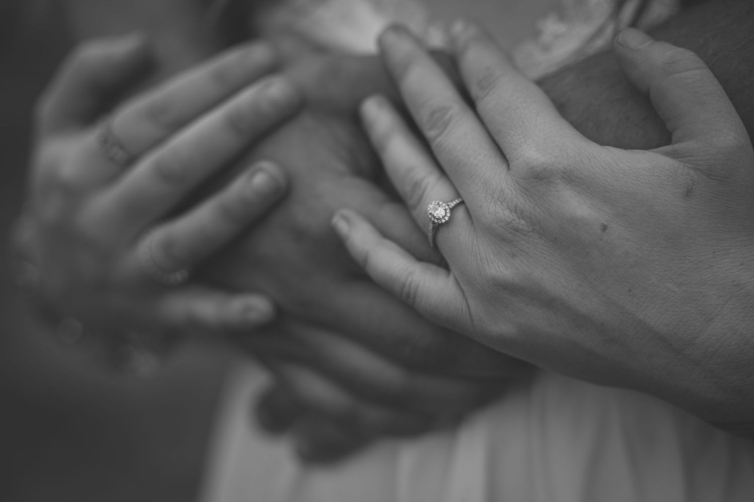 Black and white photo of two hands clasped together, one wearing a diamond engagement ring.