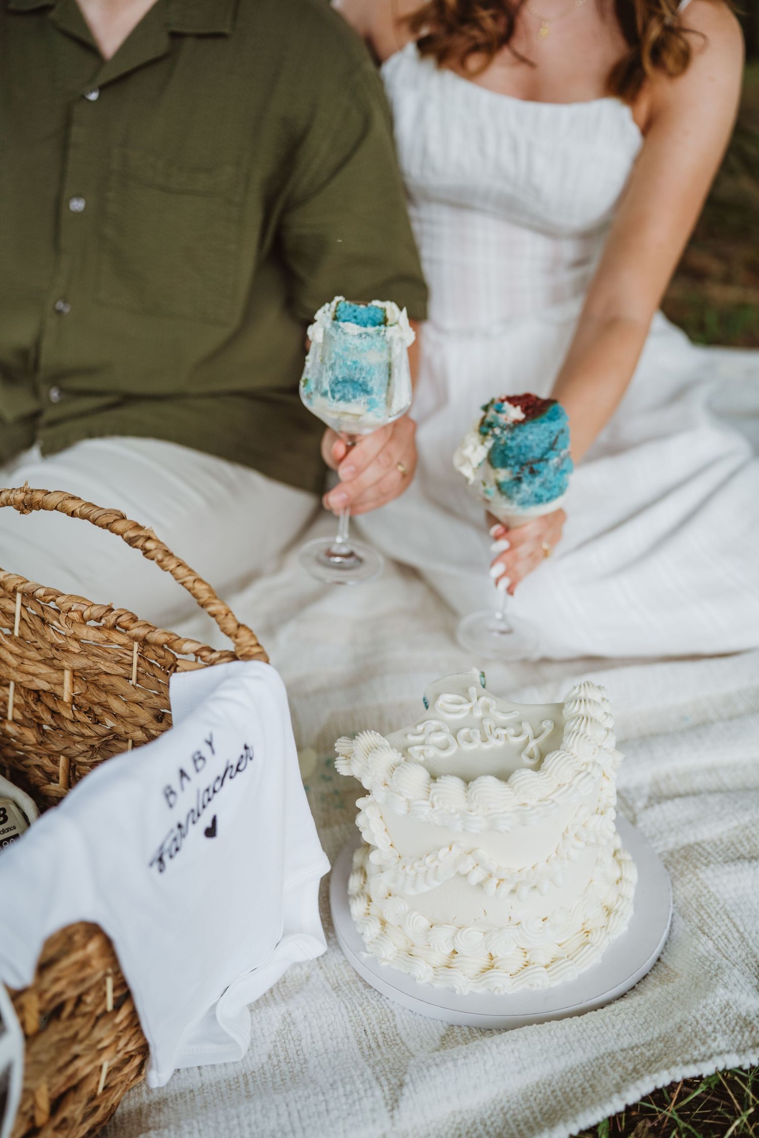 Couple sitting on picnic blanket holding blue frosted champagne glasses beside a white baby announcement cake.