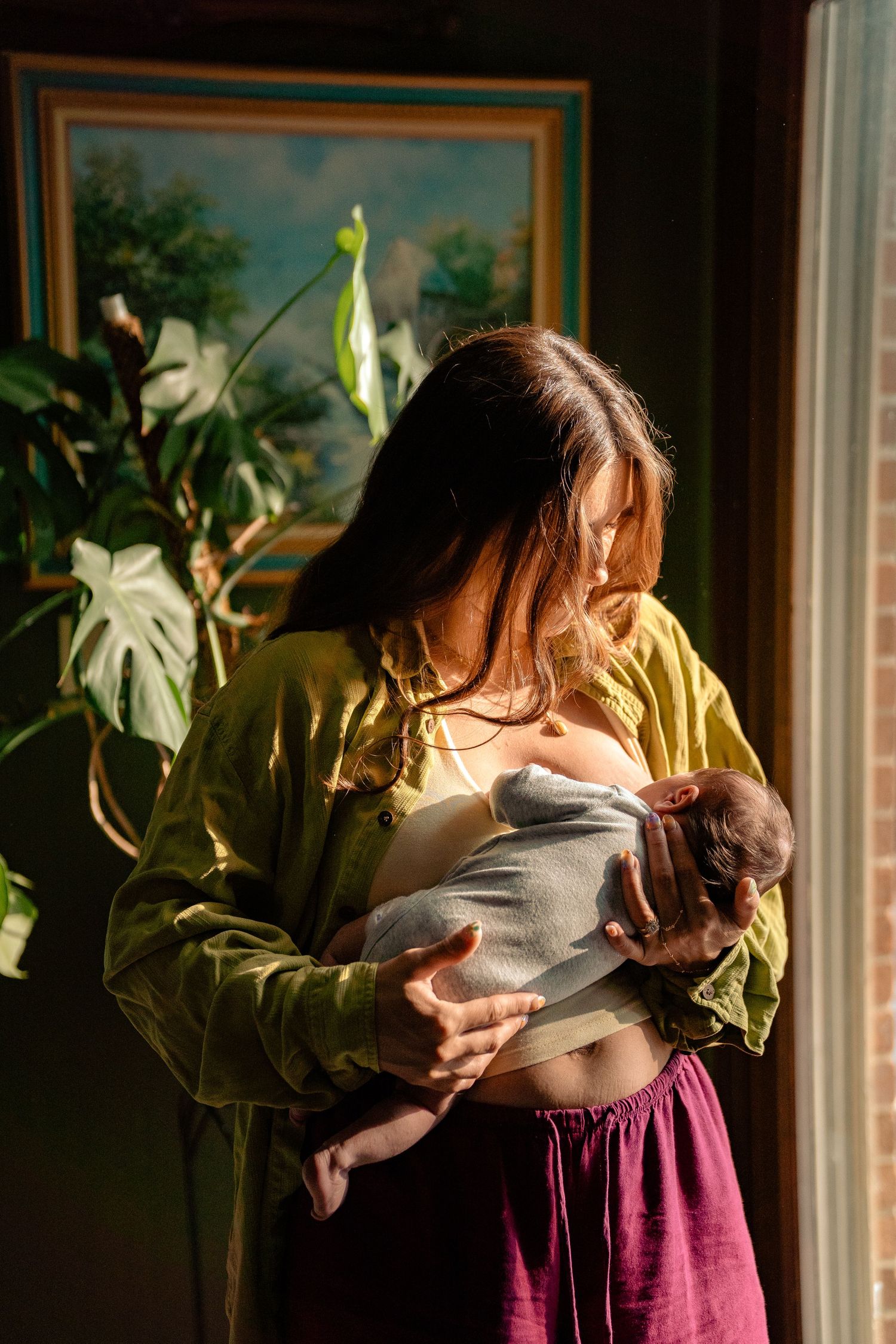 New mother in olive green shirt breastfeeding swaddled newborn by sunlit window with tropical plants.
