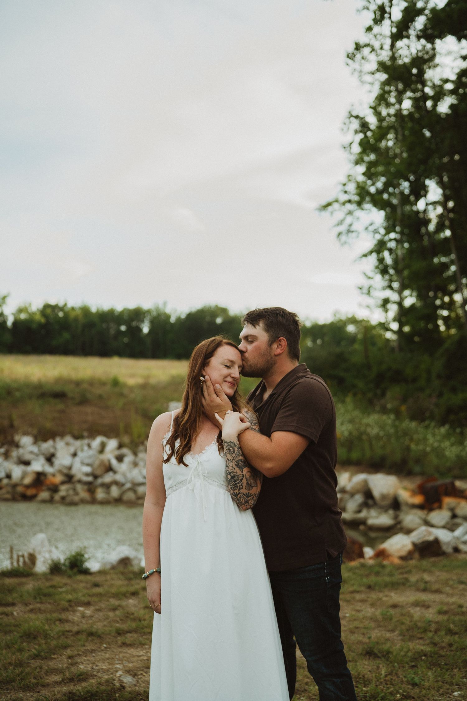 A man tenderly kisses a woman in a white dress on the cheek near a pond in a lush green field.