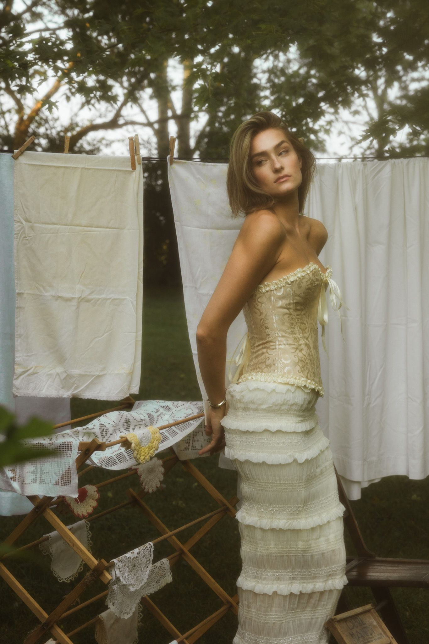 Woman in vintage white dress poses near clothesline in garden setting.