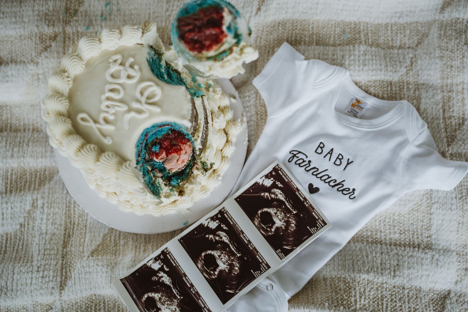 White baby announcement cake with blue floral detail beside ultrasound photos and a Baby Farbischer onesie.