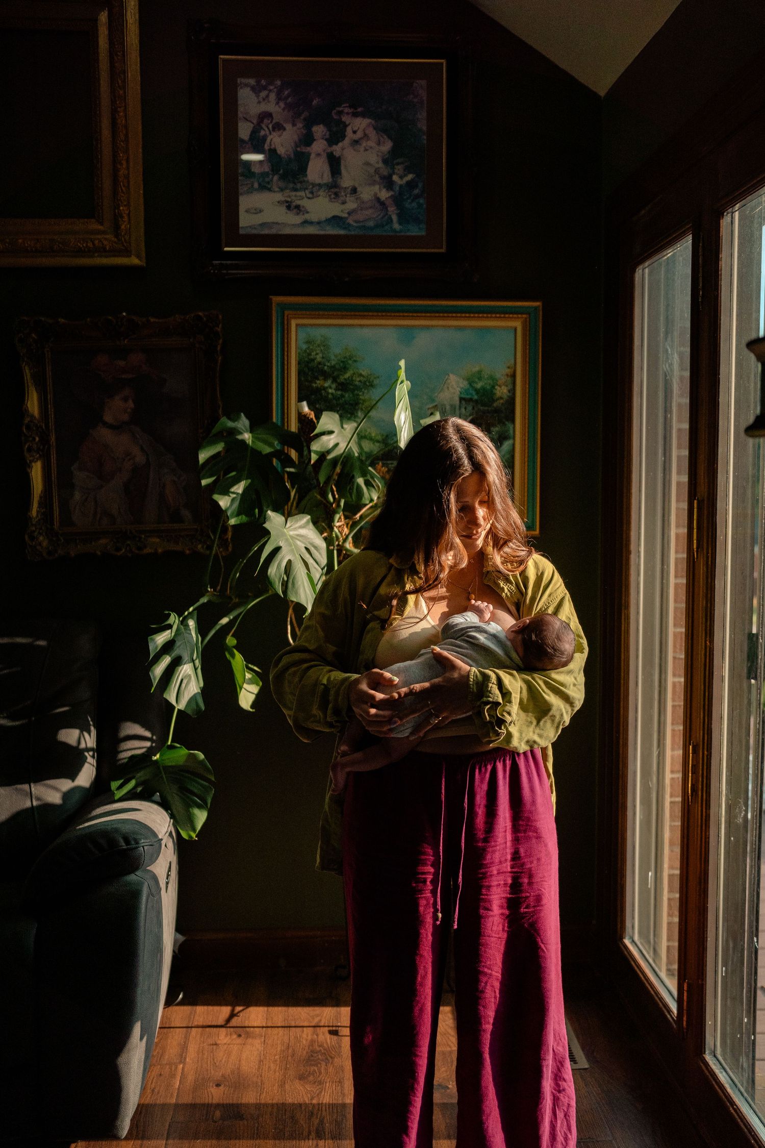 Woman in olive blouse and red pants cradles newborn baby by bright window in moody dark-walled room.