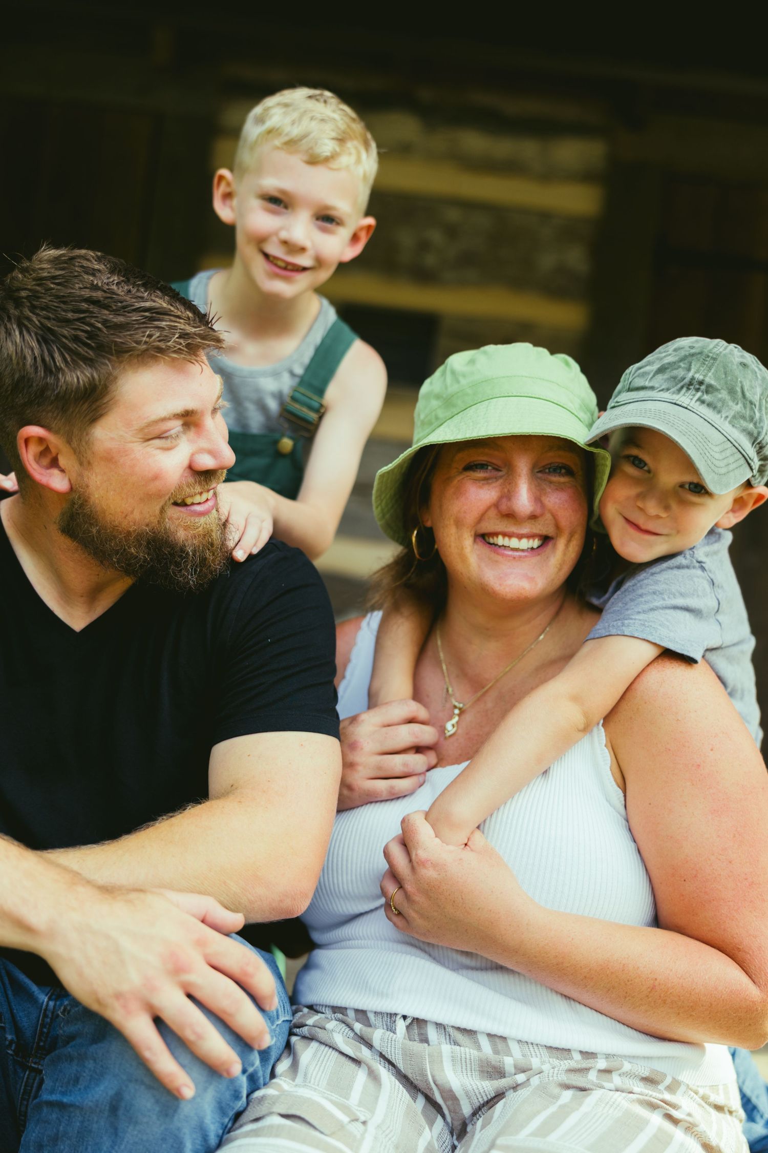 Happy family of four laughing together outdoors, with two young boys, a bearded dad, and a smiling mom in a green hat.