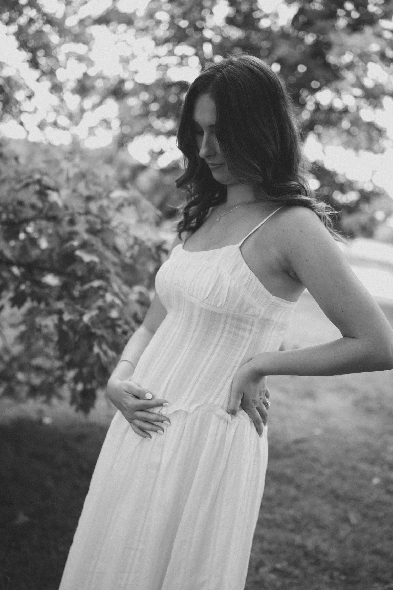 Black and white portrait of pregnant woman in white sundress cradling baby bump outdoors under trees.