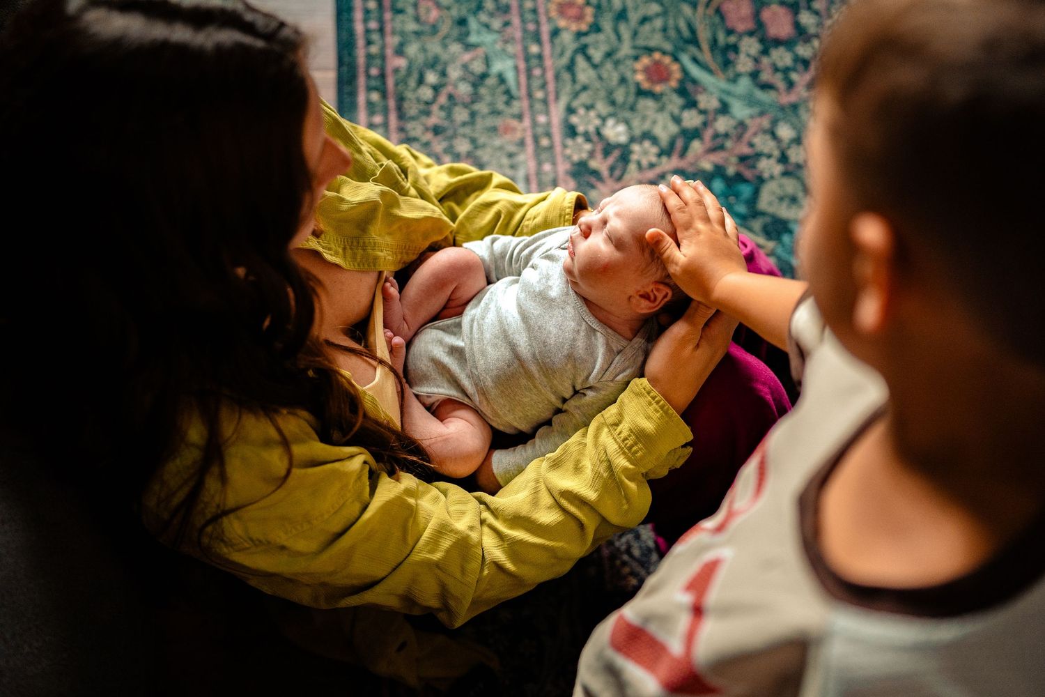 A newborn baby lying on an adult's lap while two children curiously look on in a warm family moment.