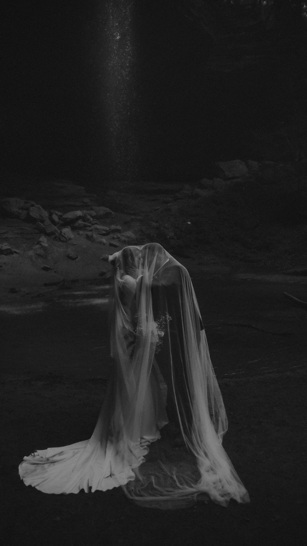 Artistic black and white wedding photo with flowing dress and veil in dark setting.