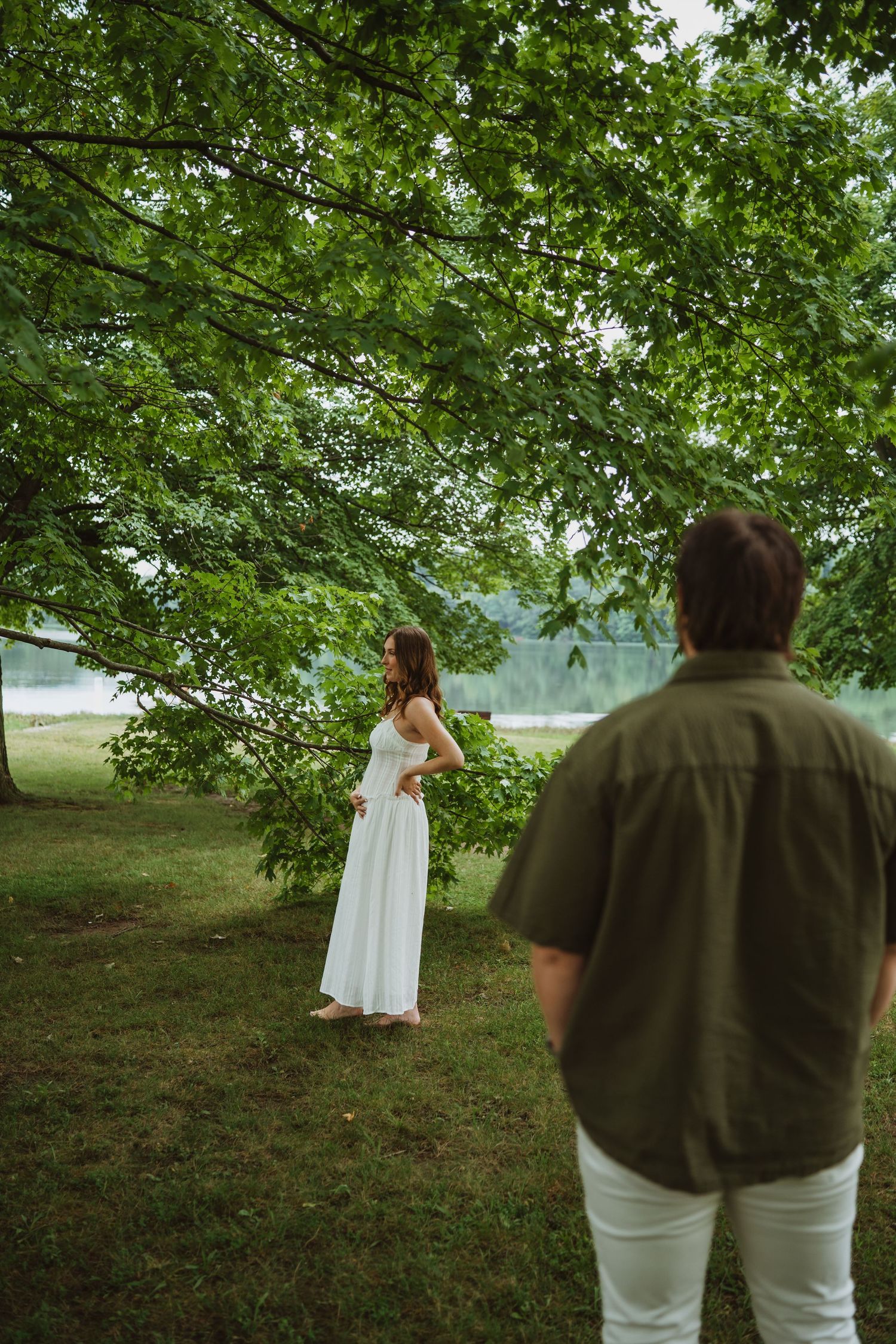 Pregnant woman in white dress stands under green tree canopy as man watches from foreground outdoors.